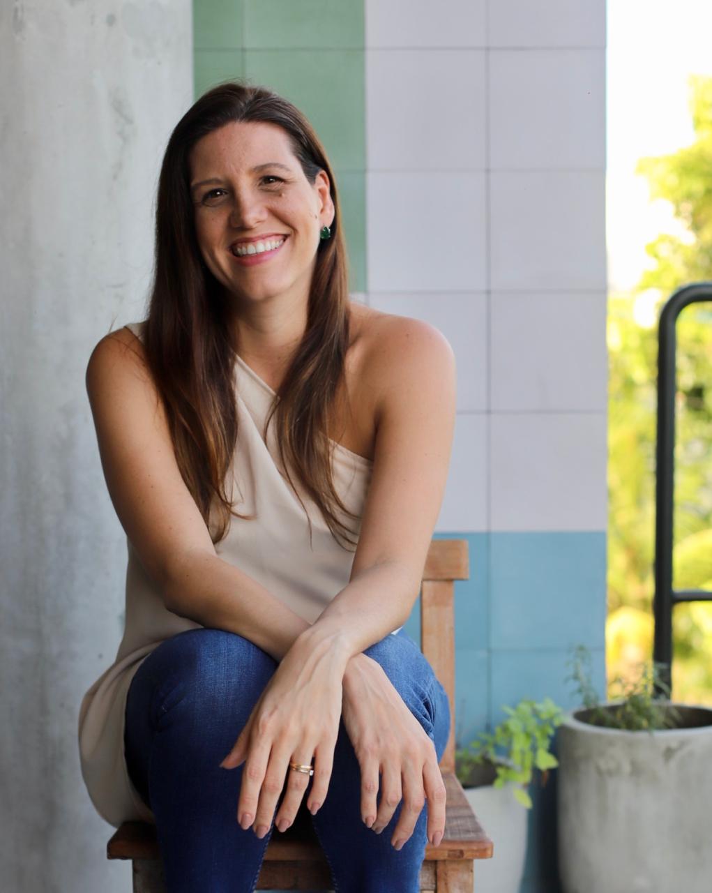 Priscilla Fontes sentada em um banco de madeira na varanda, sorrindo